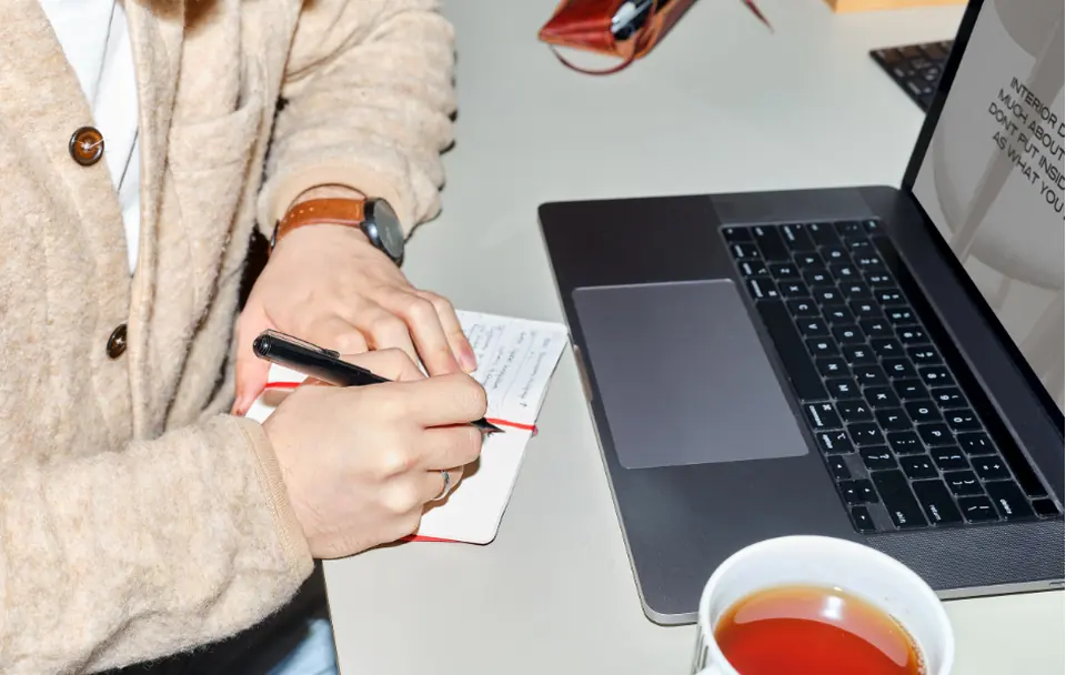 Manos escribiendo en una libreta junto a un computador y una taza de té